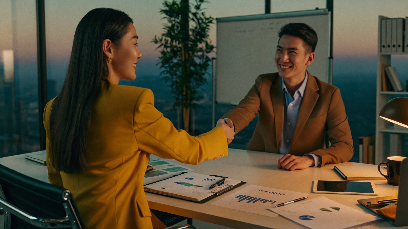 Two professionals shaking hands over a desk of performance reports and charts in a modern office at dusk